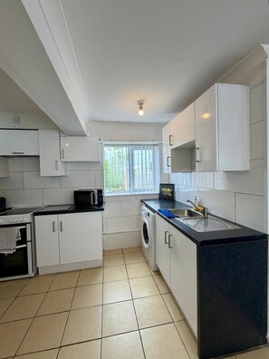 A compact kitchen with white cabinets, a microwave, a washing machine, and a sink. - 3-Bed Home, Sleeps up to 6, Free Parking Wi-Fi (Corby)