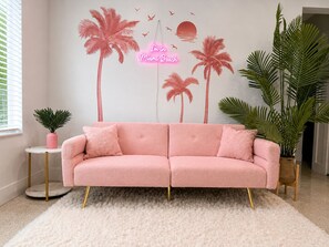 A pink sofa with gold legs, a white fluffy rug, and a wall with a palm tree mural and a neon sign. - 12 Min to Beach - Karaoke Room, Pool & Games (North Miami Beach)