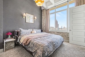 Ein Schlafzimmer mit einem großen Bett, einem Nachttisch mit Blumenvase, Blick auf die Stadt durch das Fenster und einer Tür rechts.