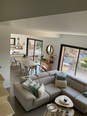 Un salon moderne avec un canapé d’angle, un coin repas doté d’une table en bois et de grandes fenêtres offrant une vue sur l’extérieur.