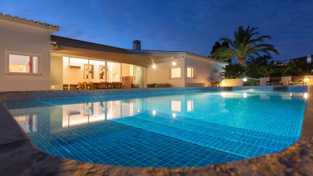 En swimmingpool med flisegulv, en overdækket patio med siddepladser, og en bygning med vinduer og døre.