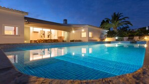 Une piscine avec un revêtement de sol en carrelage, un patio couvert avec des sièges, et un bâtiment doté de fenêtres et de portes.