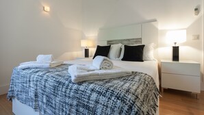 Et hotelværelse med en pænt redt seng, et sengebord med en lampe og en vægmonteret lampe.