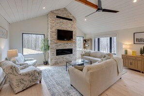A spacious living room with a stone fireplace, a ceiling fan, and large windows offering a view of the outdoors. - Modern Mountain Retreat at Waterville Estates (Campton)