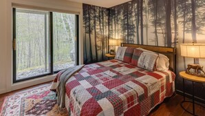 Ein Schlafzimmer mit einem großen Bett, einem Wandgemälde zum Thema Wald, einem Fenster mit Blick auf Bäume und einem kleinen runden Tisch mit Lampe.