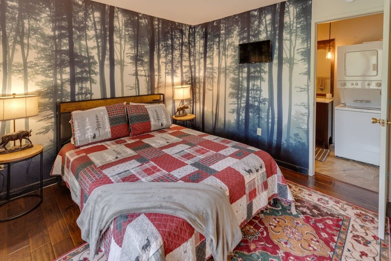 Ein Schlafzimmer mit einem großen Bett, einer Wand mit Waldmotiv, einem Fernseher und einer Küchenzeile.