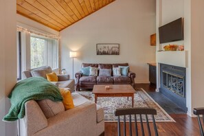 Een woonkamer met een bank, fauteuil, salontafel, open haard en een aan de muur bevestigde televisie.