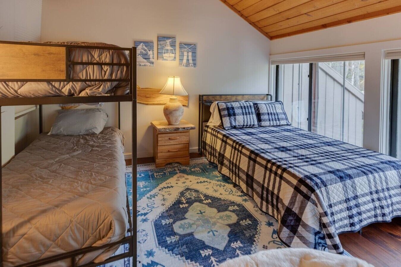 Een slaapkamer met een stapelbed, houten plafond, nachtkastje met lamp, getextileerd vloerkleed en bed met ruitdessin.
