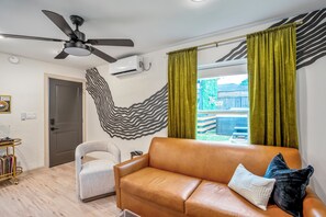 Una sala de estar con un sofá de cuero marrón, un sillón blanco, un ventilador de techo y vista a un patio trasero a través de una ventana con cortinas verdes.