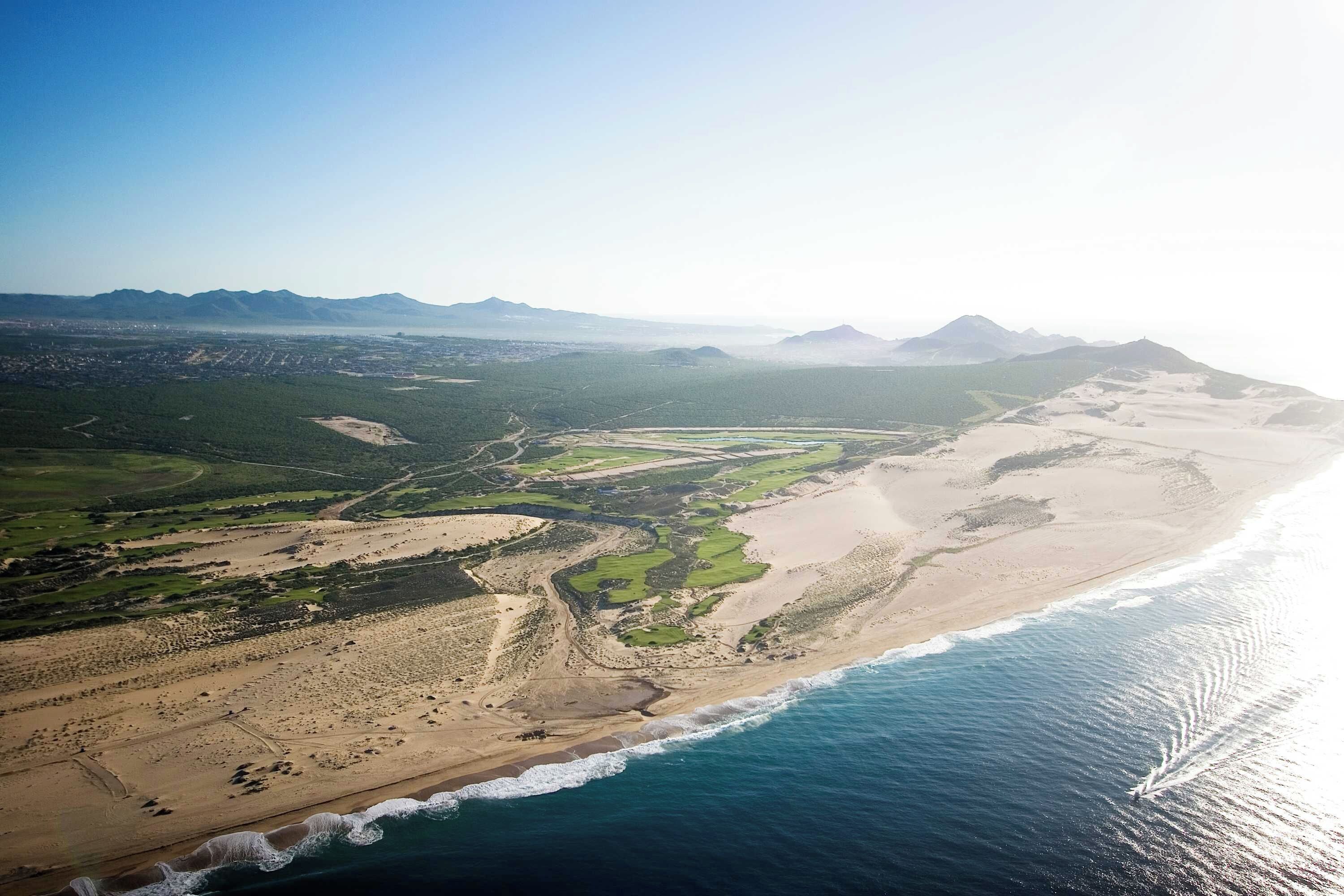 An aerial view of a coastal landscape with a beach, green fields, and distant mountains.