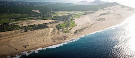 An aerial view of a coastal landscape with a beach, green fields, and distant mountains.