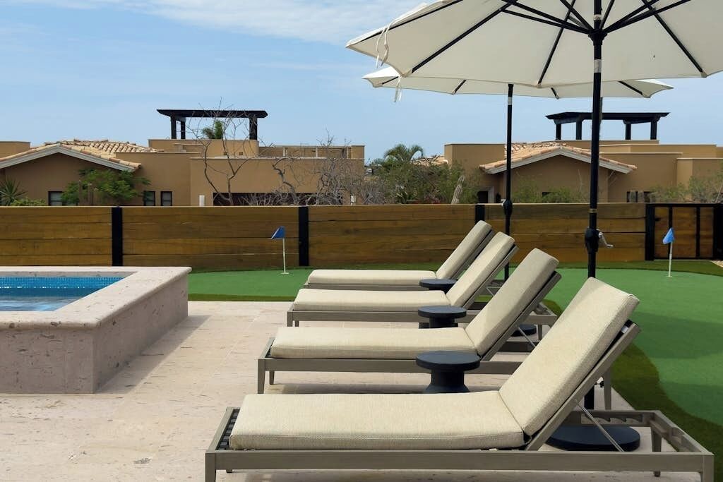 A poolside area with lounge chairs and an umbrella, a pool, and a wooden fence.