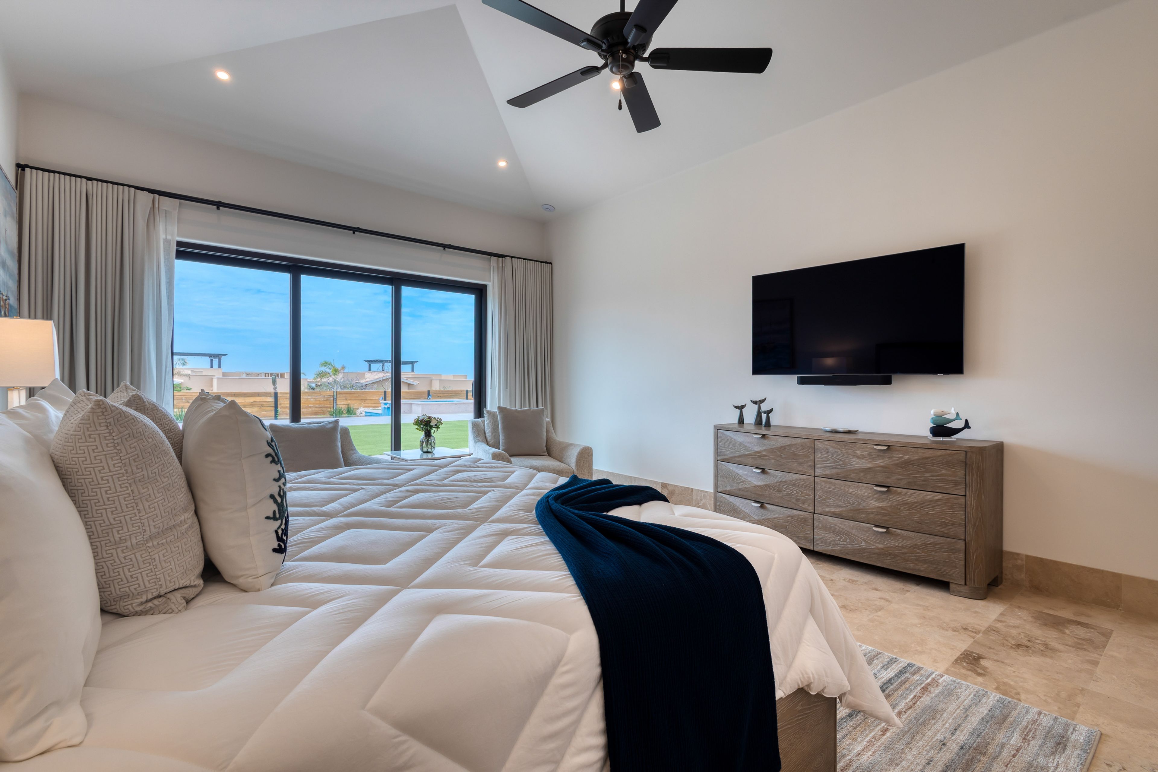 A bedroom with a large bed, a ceiling fan, a TV, a wooden dresser, and a view of the outdoors.