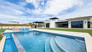En swimmingpool med en patio, et overdækket loungeområde og et moderne hus med store vinduer.