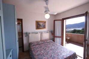 A bedroom with a bed, a ceiling fan, a painting, and a balcony with a view. - Le Corti Di Marinella 6p Garden View (Marinella)