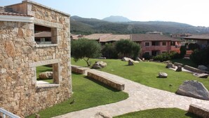 A stone building with a stone pathway, a grassy area with rocks, and a view of other buildings in the background. - Le Corti Di Marinella 4p Garden View (Marinella)