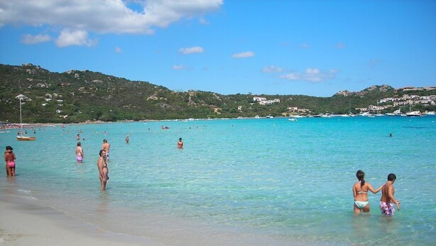 A beach with clear turquoise water, people in the water, and buildings on the hillside in the background. - Le Corti Di Marinella 4p Garden View (Marinella)