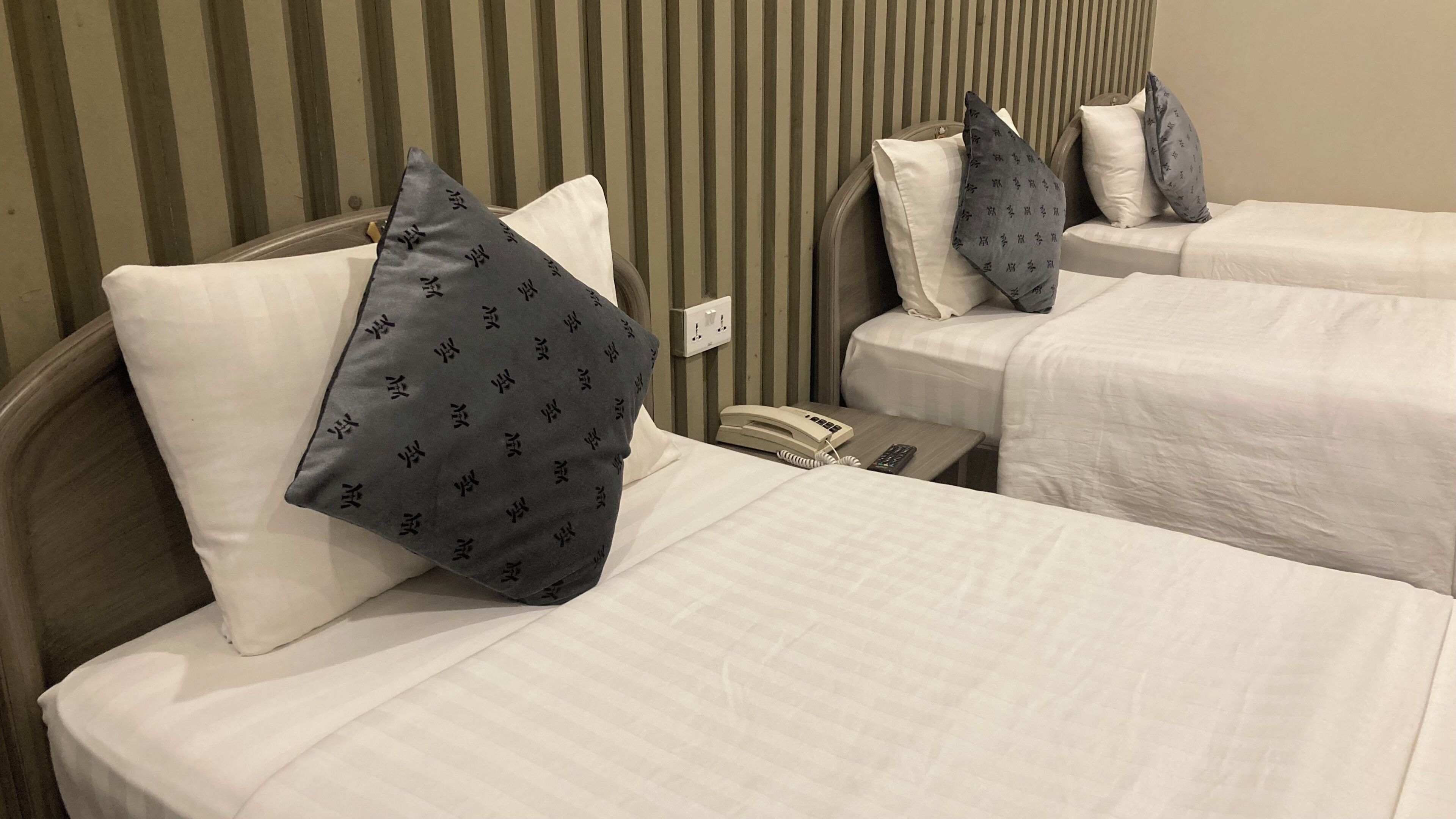 A hotel room with two beds, each with white bedding and patterned pillows. There is a bedside table with a telephone and a remote control.