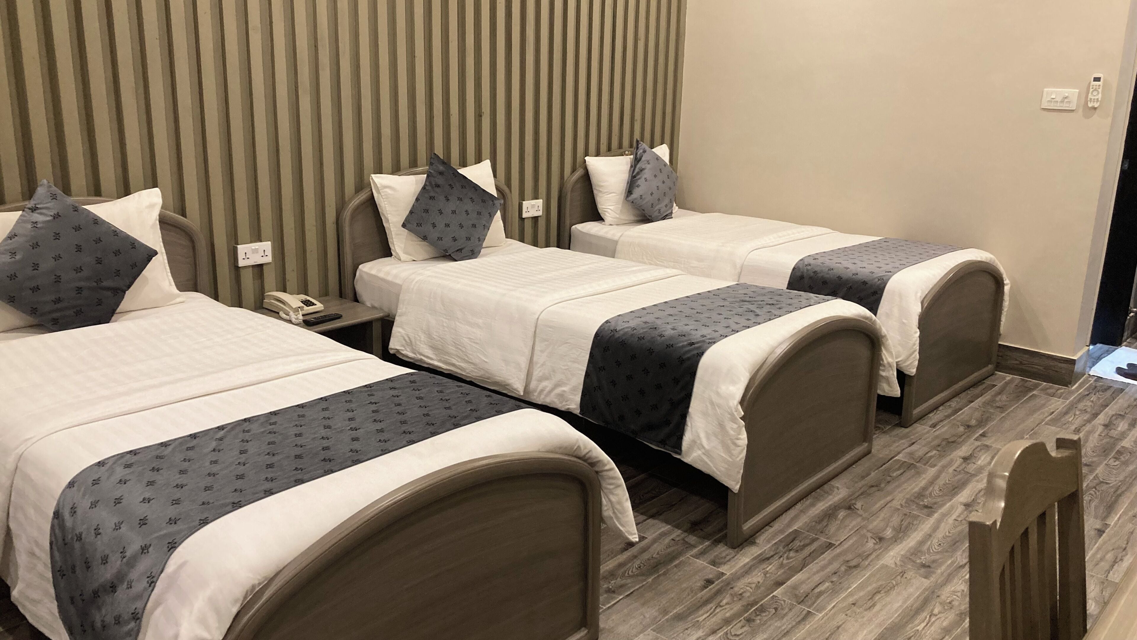 A hotel room with three single beds, each with a pillow and a blanket. There is a bedside table with a phone and a lamp. The room has a striped wallpaper and a wooden floor.