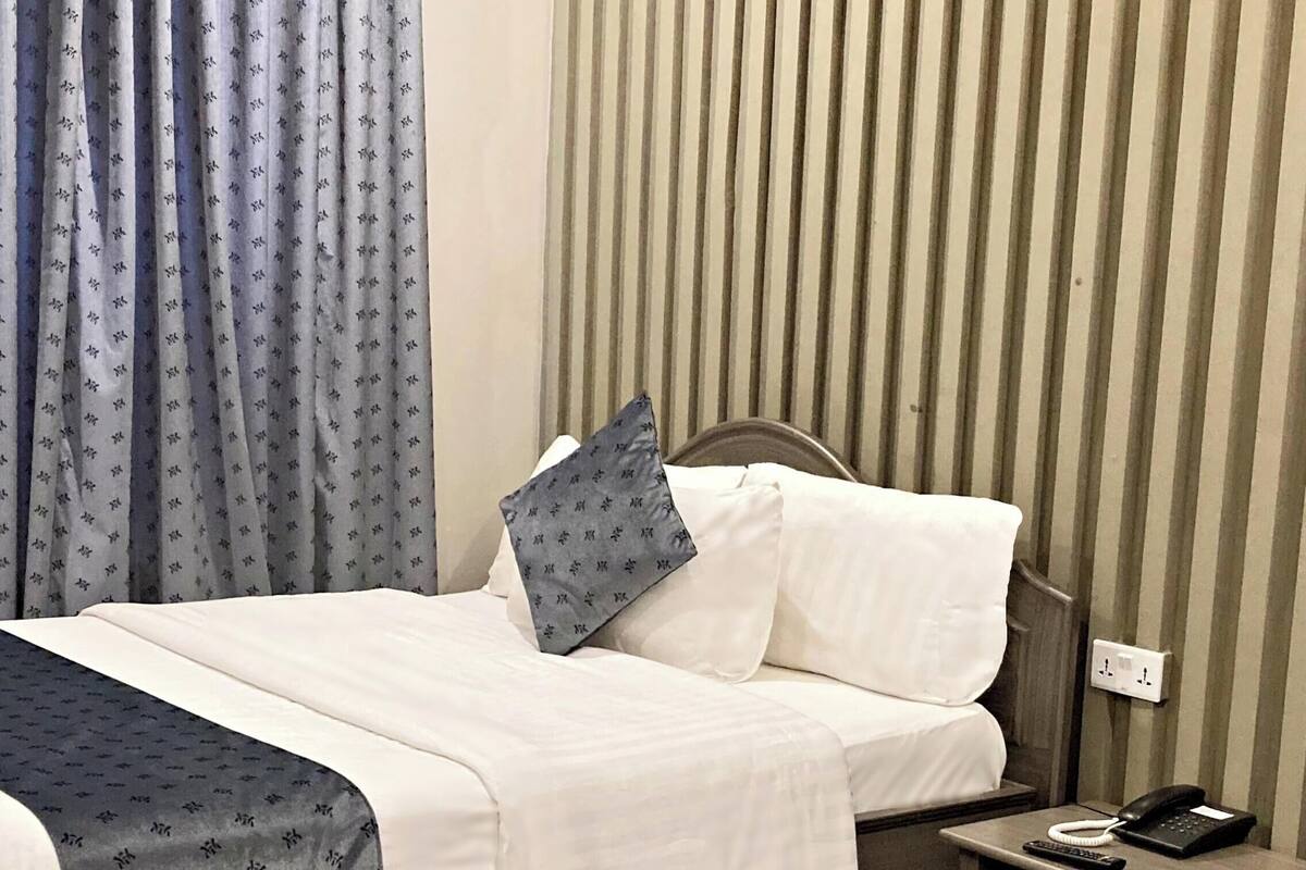 Una habitación de hotel con cama, mesita de noche, teléfono, control remoto, almohada y cortinas.
