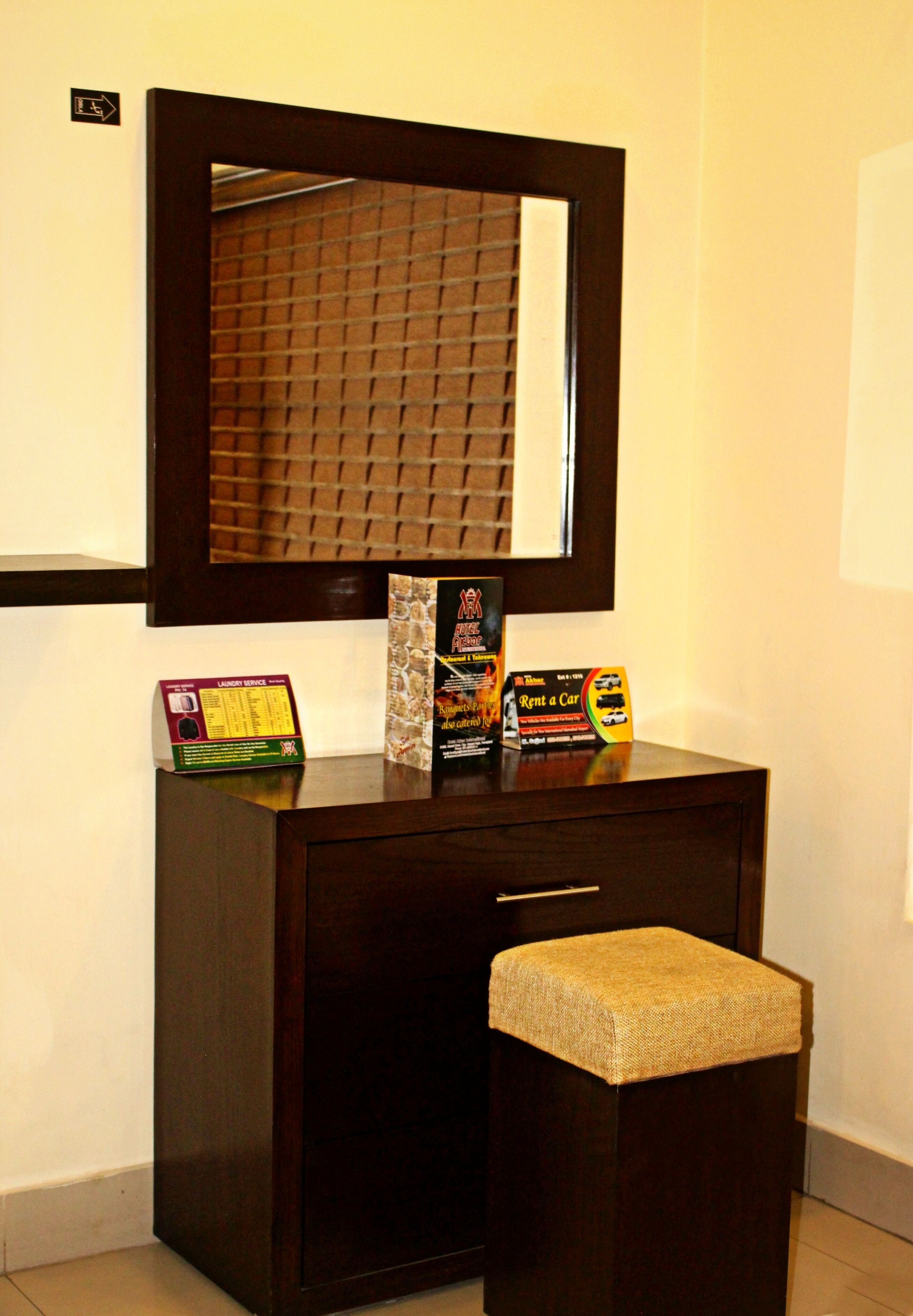 A dark wooden vanity with a mirror, a small stool, and a few items on top including a box labeled 'Laundry Service' and another box with a 'Rent a Car' sign.