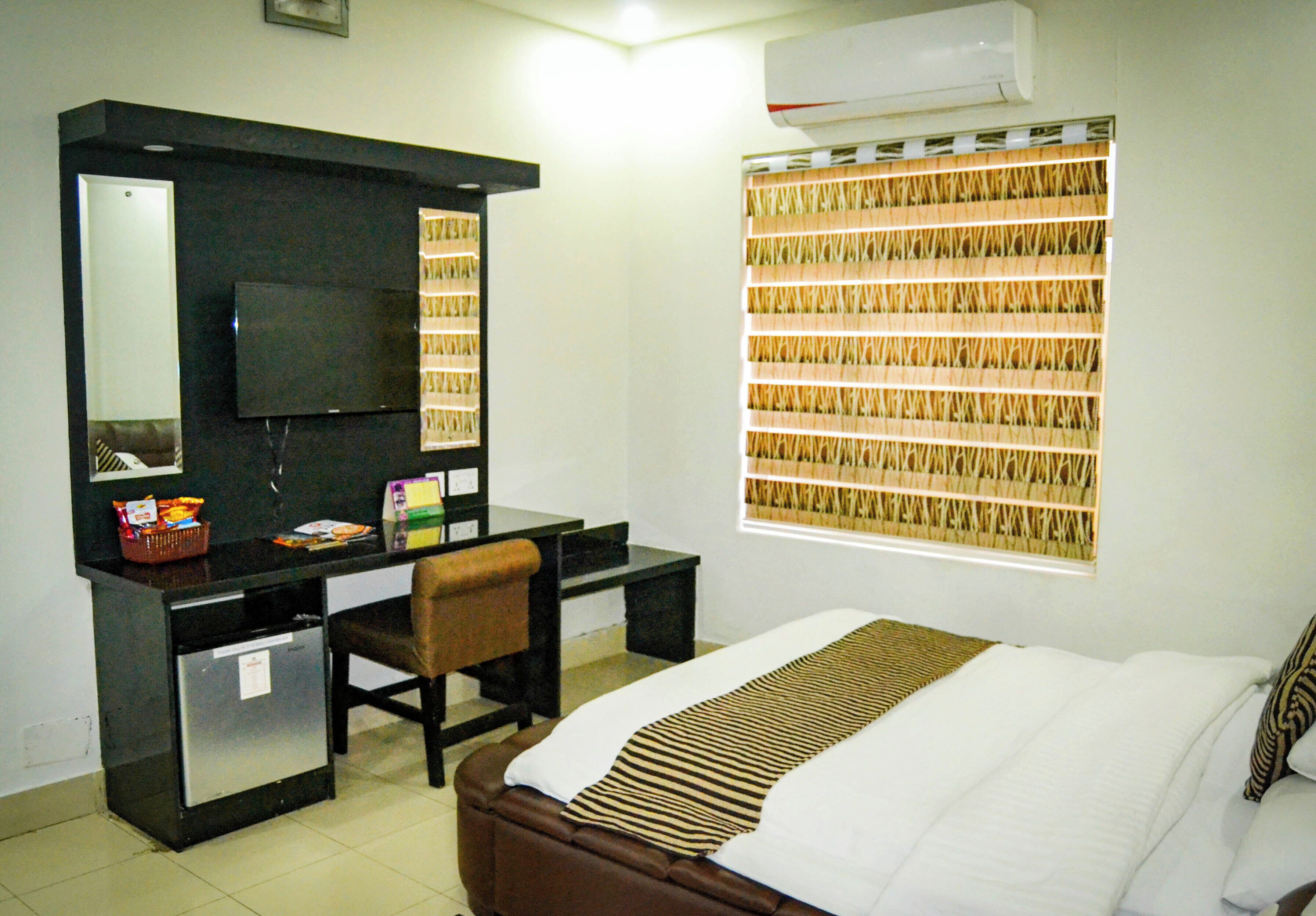 Habitación de hotel con cama, escritorio con silla, televisor, refrigerador pequeño y ventana con persiana tejida.