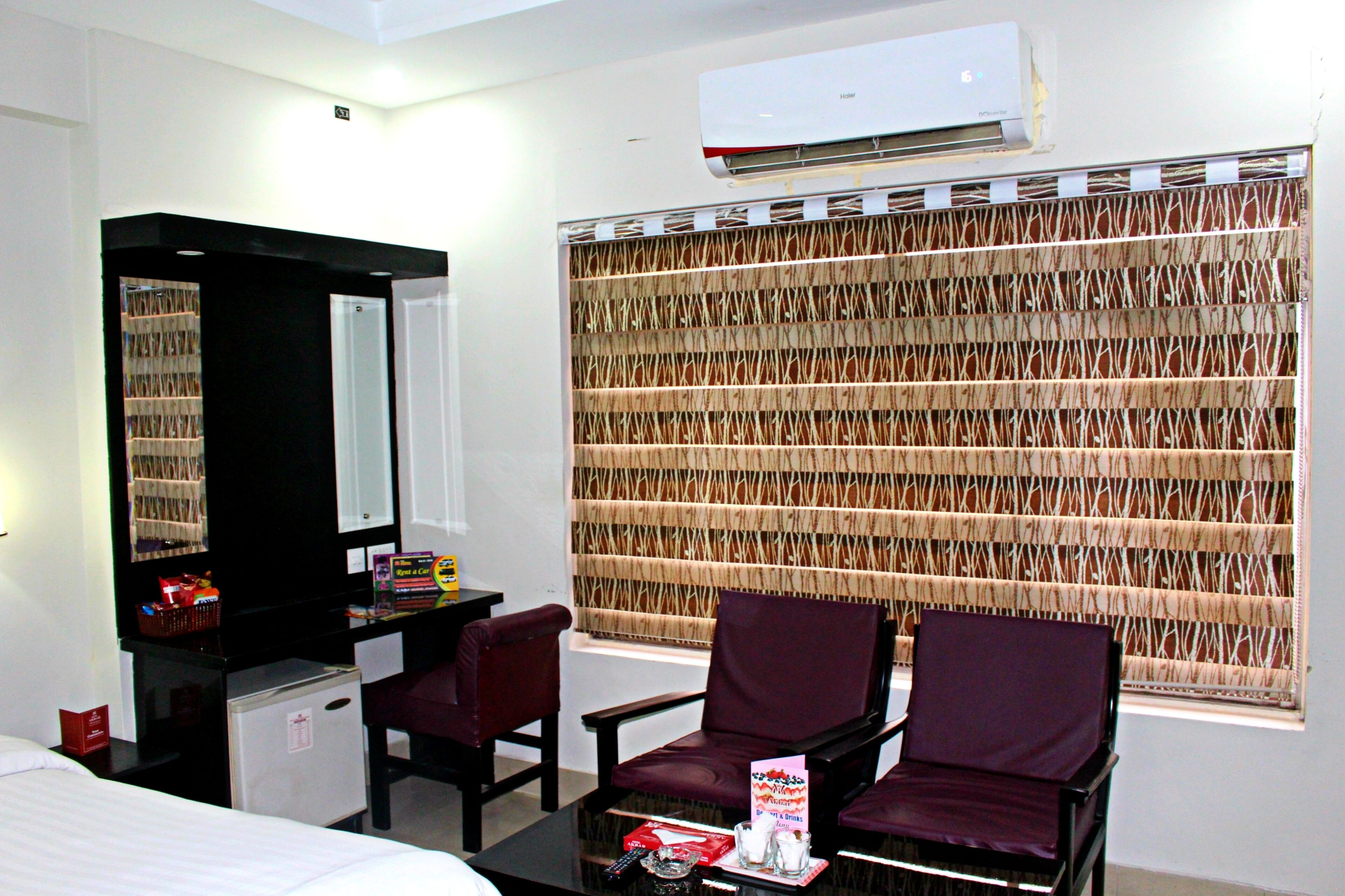 A hotel room with a bed, a desk with a mirror, two chairs, a small refrigerator, and a window with a patterned blind.
