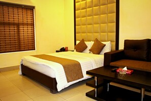 A hotel room with a bed, a sofa, a coffee table, and a window with blinds. - Hotel Akbar International (Rawalpindi)