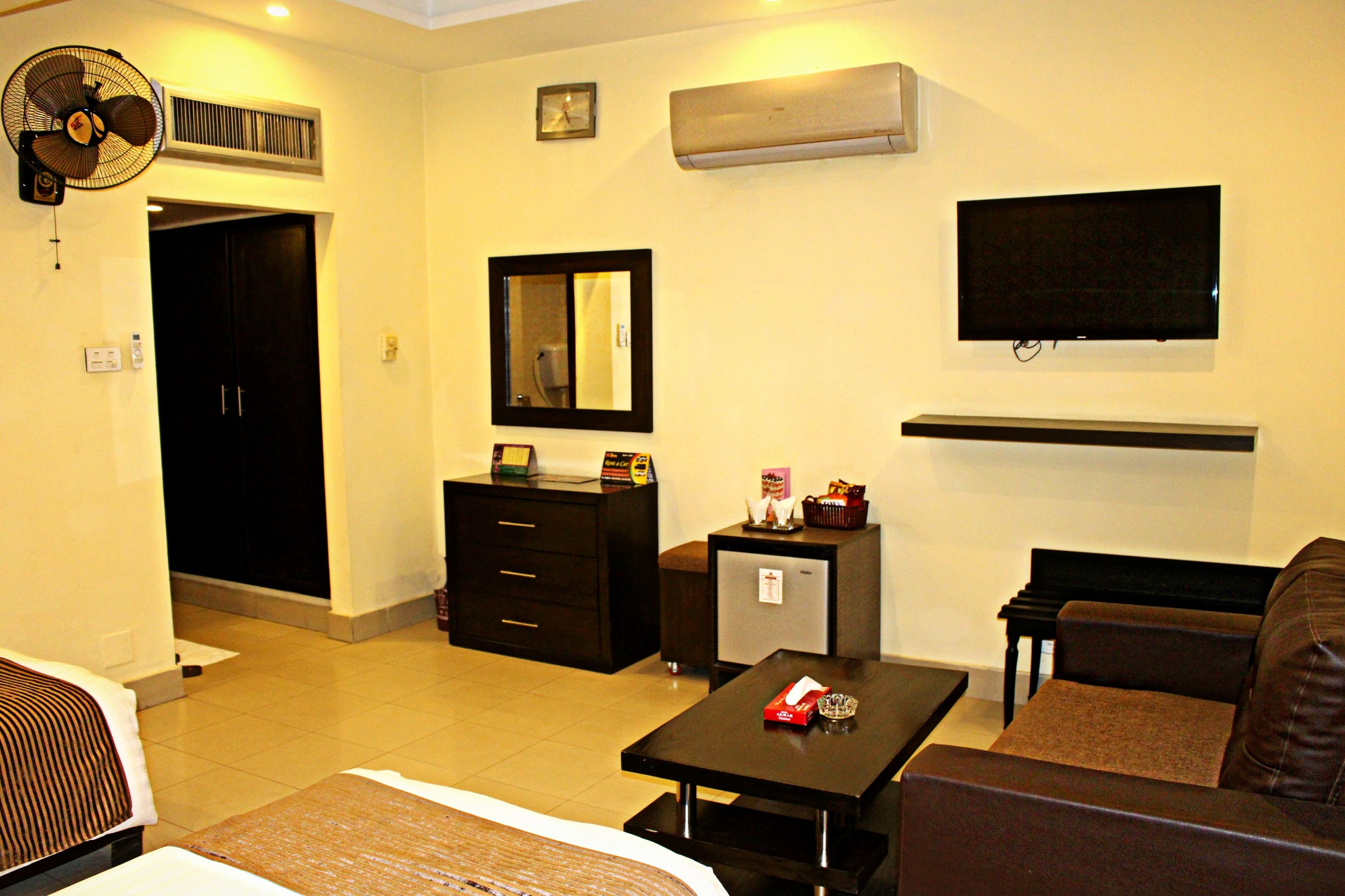 A hotel room with a bed, a desk, a chair, a small refrigerator, a mirror, an air conditioning unit, a television, and a fan.