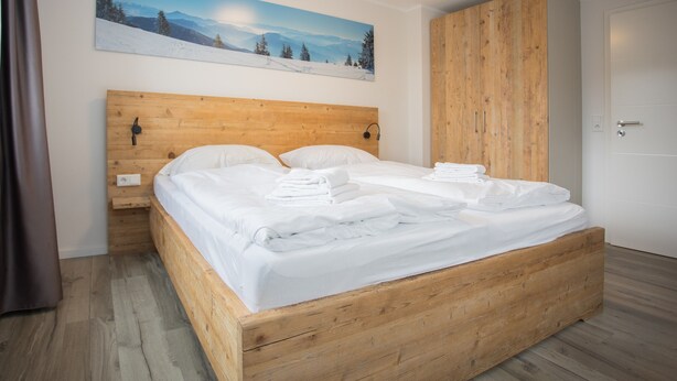 A bedroom with a wooden bed, white linens, a wooden headboard, a wooden wardrobe, and a painting of a snowy landscape. - Appartement - Winterbergerstrasse 3, nr. 511 | Neuastenberg (Winterberg)