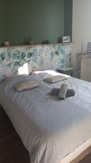 Uma cama bem arrumada com almofadas e um cobertor, uma estante com objetos decorativos e uma parede com padrão de folhas.