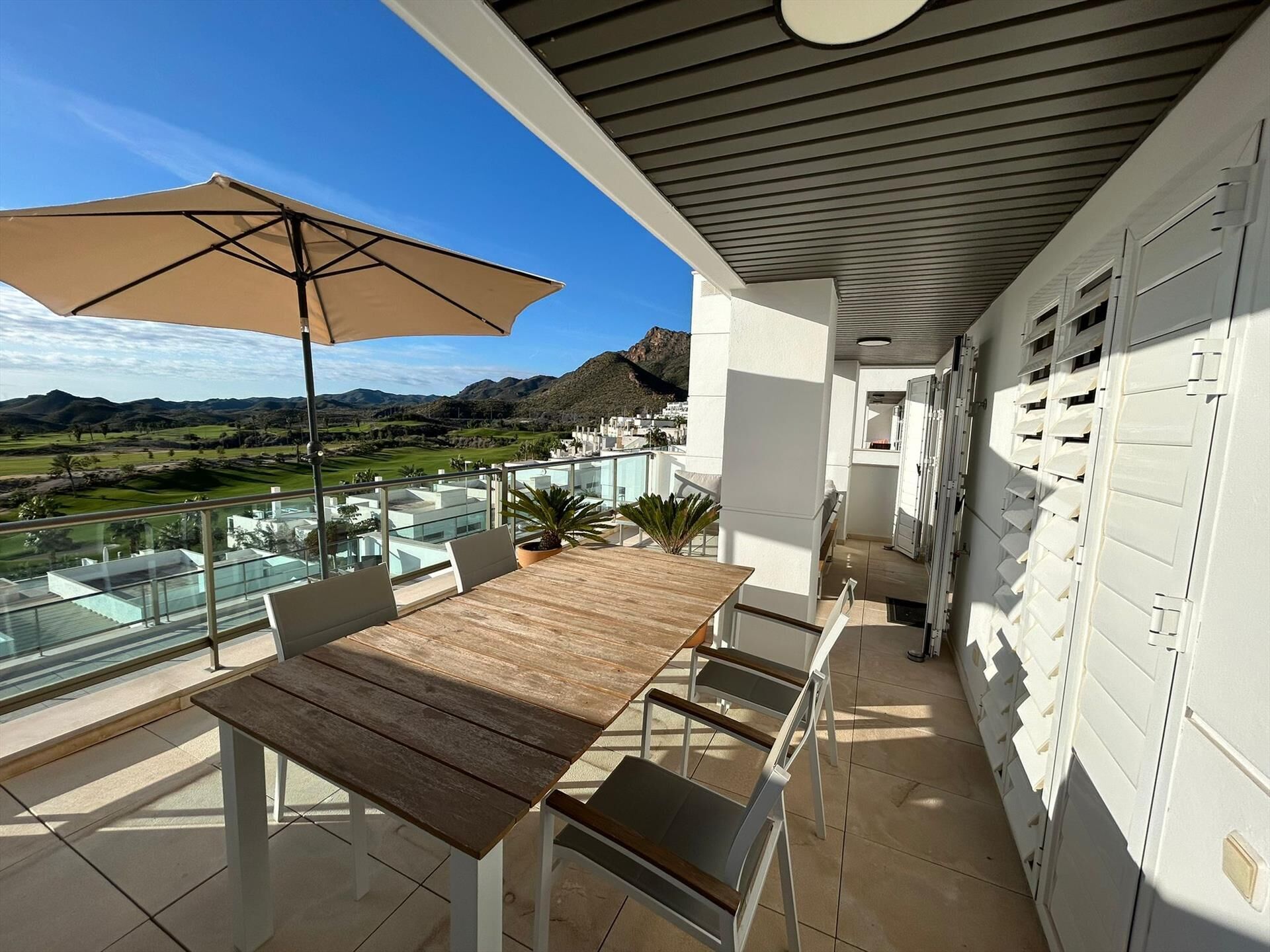 A balcony with a table and chairs, a large umbrella, and a view of a golf course and mountains.