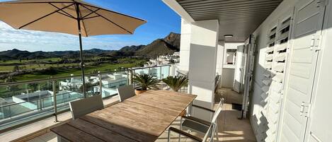 A balcony with a table and chairs, a large umbrella, and a view of a golf course and mountains.