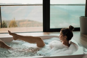 Una persona relajándose en un jacuzzi con vistas a montañas a través de un ventanal amplio.