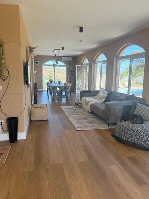 Ein großzügiger Wohnbereich mit Holzboden, einem grauen Sofa, einem Esstisch mit Stühlen und großen Bogenfenstern mit Blick ins Freie.