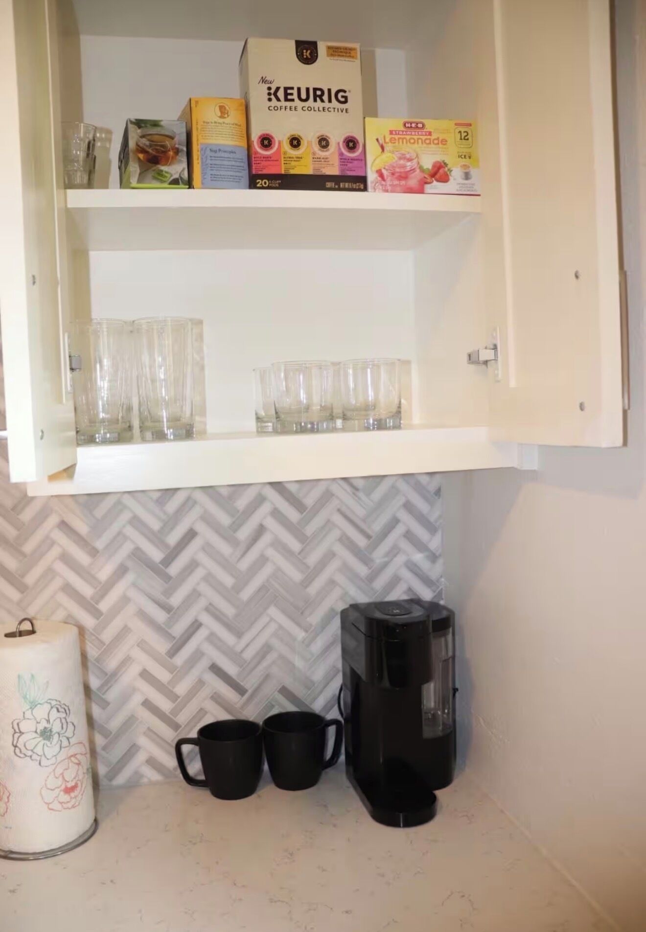 A kitchen with a coffee maker, two black mugs, and a box of Keurig coffee pods on the counter.
