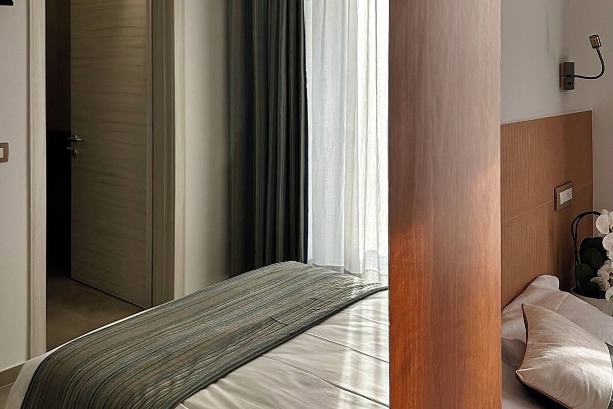 Habitación de hotel con cama, cabecera de madera, mesita de noche con lámpara y puerta que da a otra habitación.