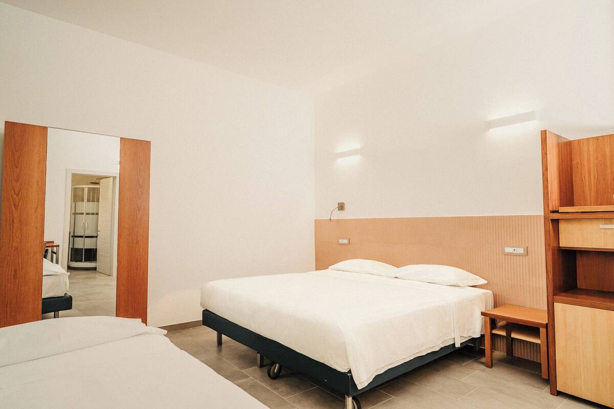 Habitación de hotel con una cama, una mesita de noche de madera, un espejo y una puerta que da a un baño.