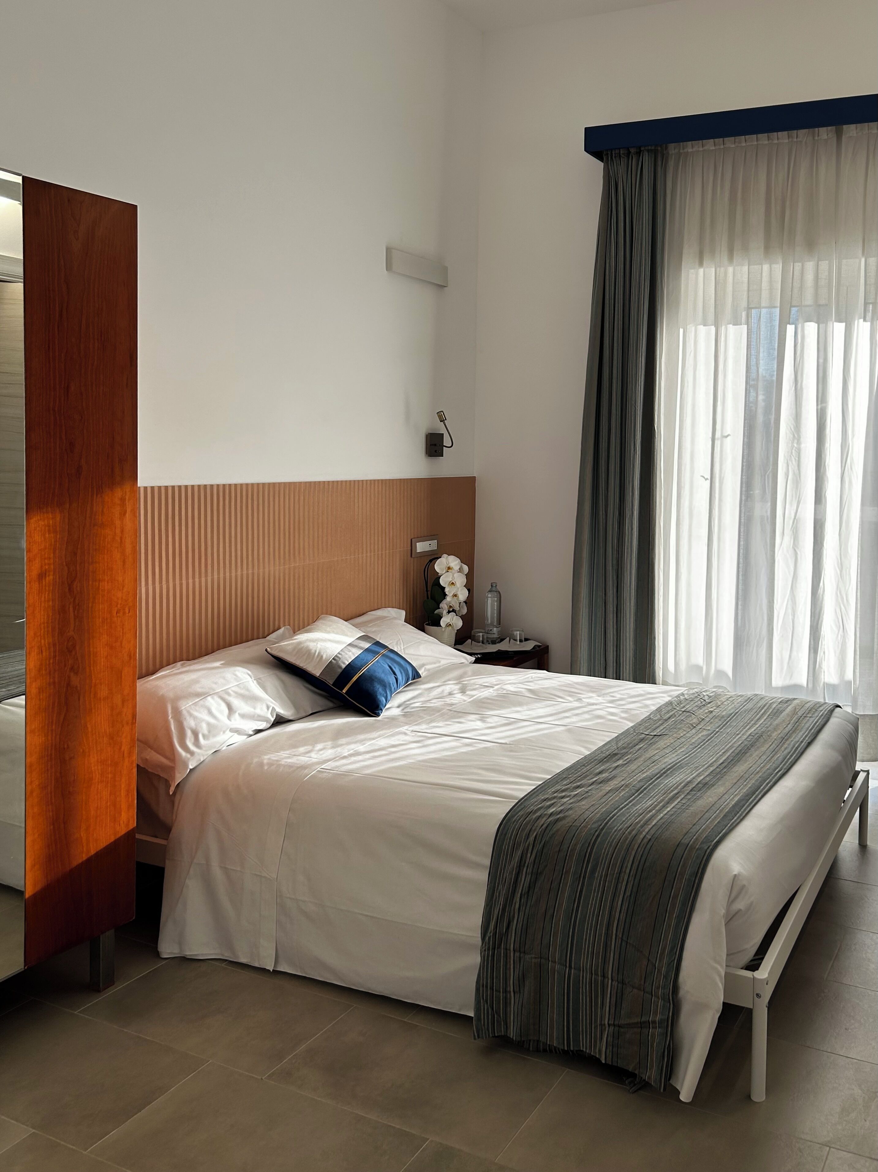 A neatly made bed with white bedding and a blue accent pillow, a bedside table with a lamp, and a window with sheer curtains.