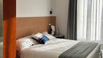 A neatly made bed with white bedding and a blue accent pillow, a bedside table with a lamp, and a window with sheer curtains.