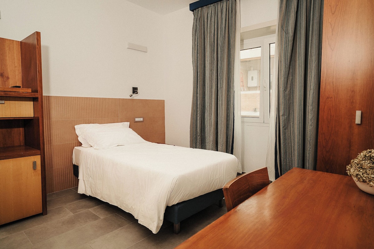 Una cama individual con cabecera de madera, una mesa de madera y un armario de madera en una habitación con ventana y cortinas.