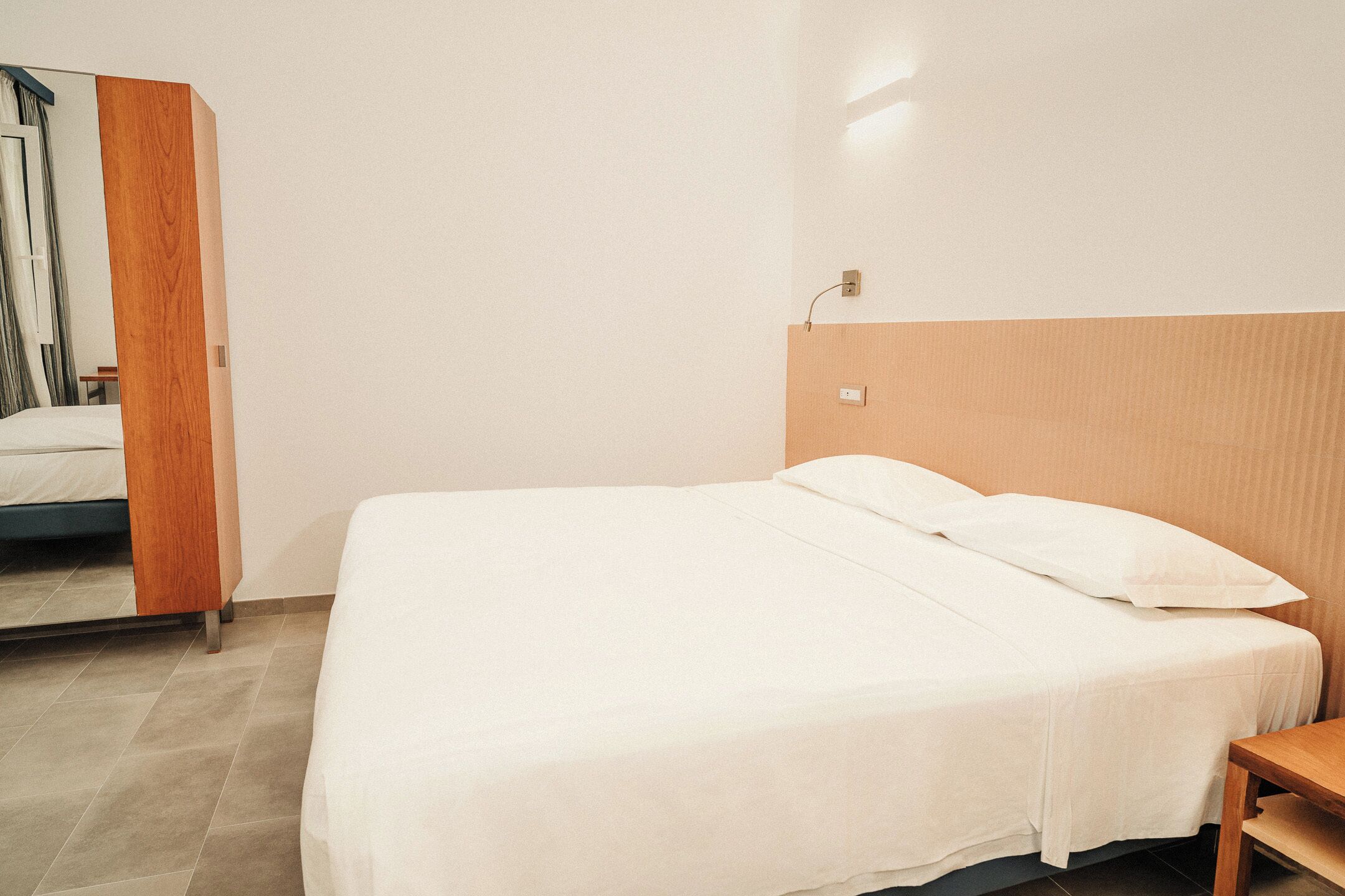 Una cama individual con cabecera de madera en una habitación con paredes lisas y suelo de baldosas.