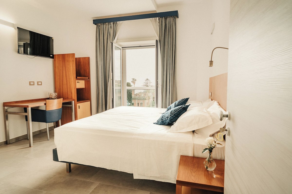 Habitación de hotel con cama, escritorio, televisor y vistas al exterior.
