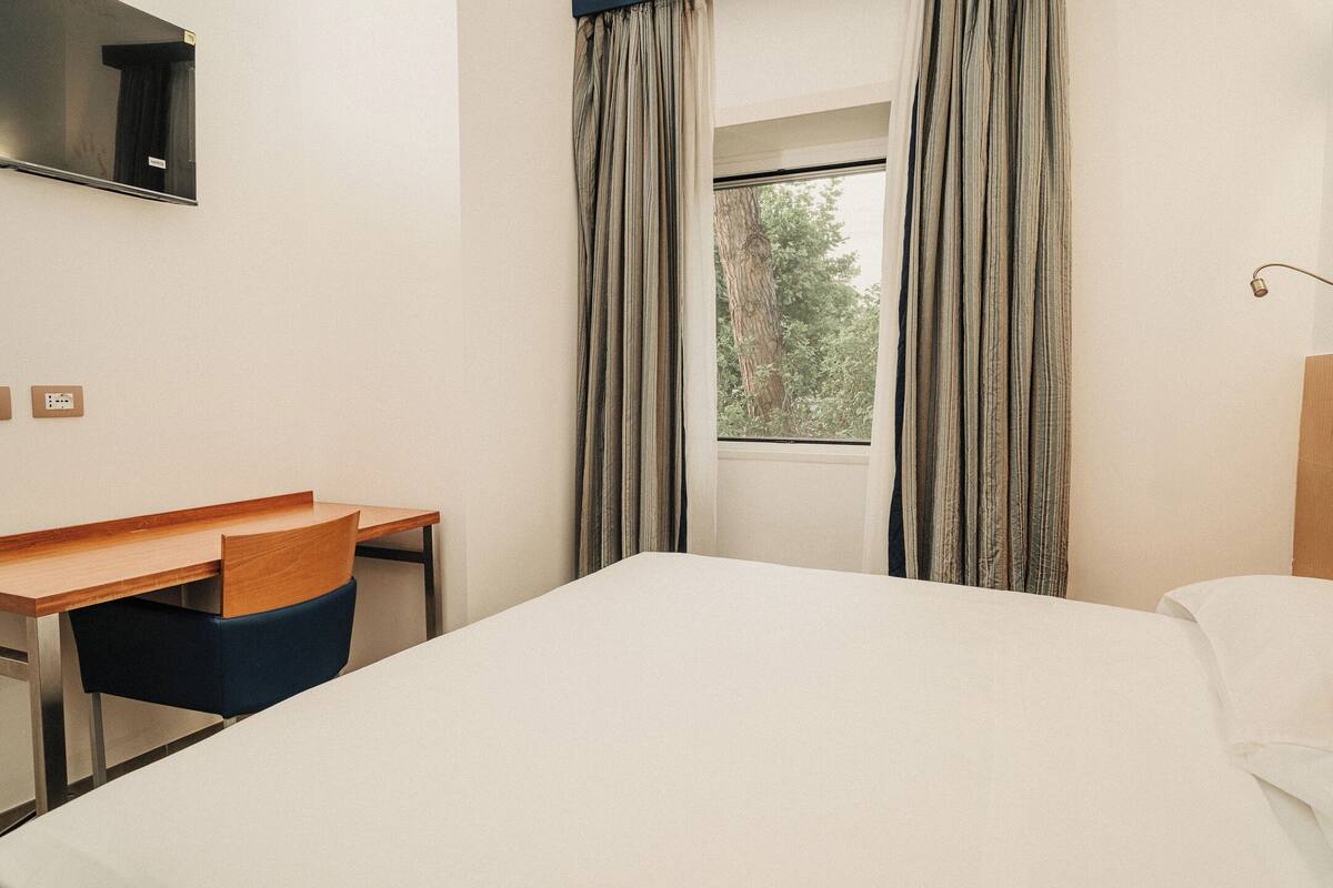 Habitación de hotel con cama, escritorio, silla, televisor montado en la pared y ventana con cortinas.