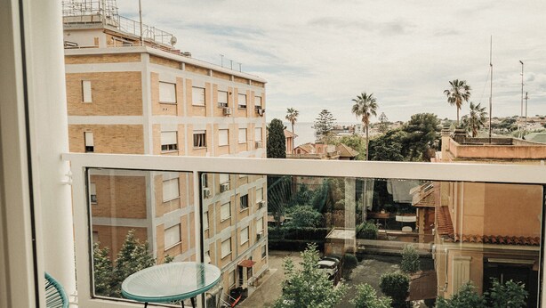 A balcony with a glass table and chairs, overlooking a residential area with buildings and trees. - Hotel Mediterraneo (Santa Marinella)