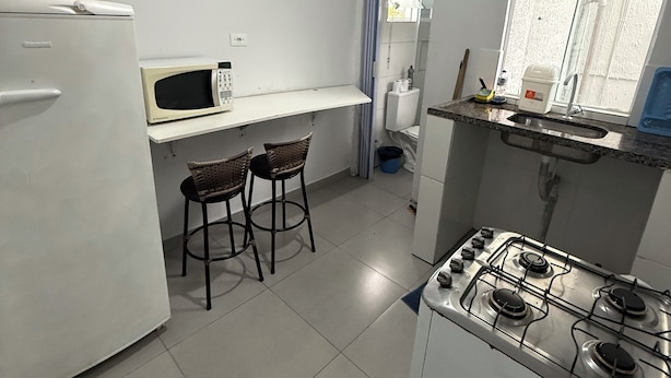 A compact kitchen with a refrigerator, microwave, gas stove, two bar stools, a window, and a sink. - Furnished studio apartment in Vila Amália, North Zone (São Paulo)