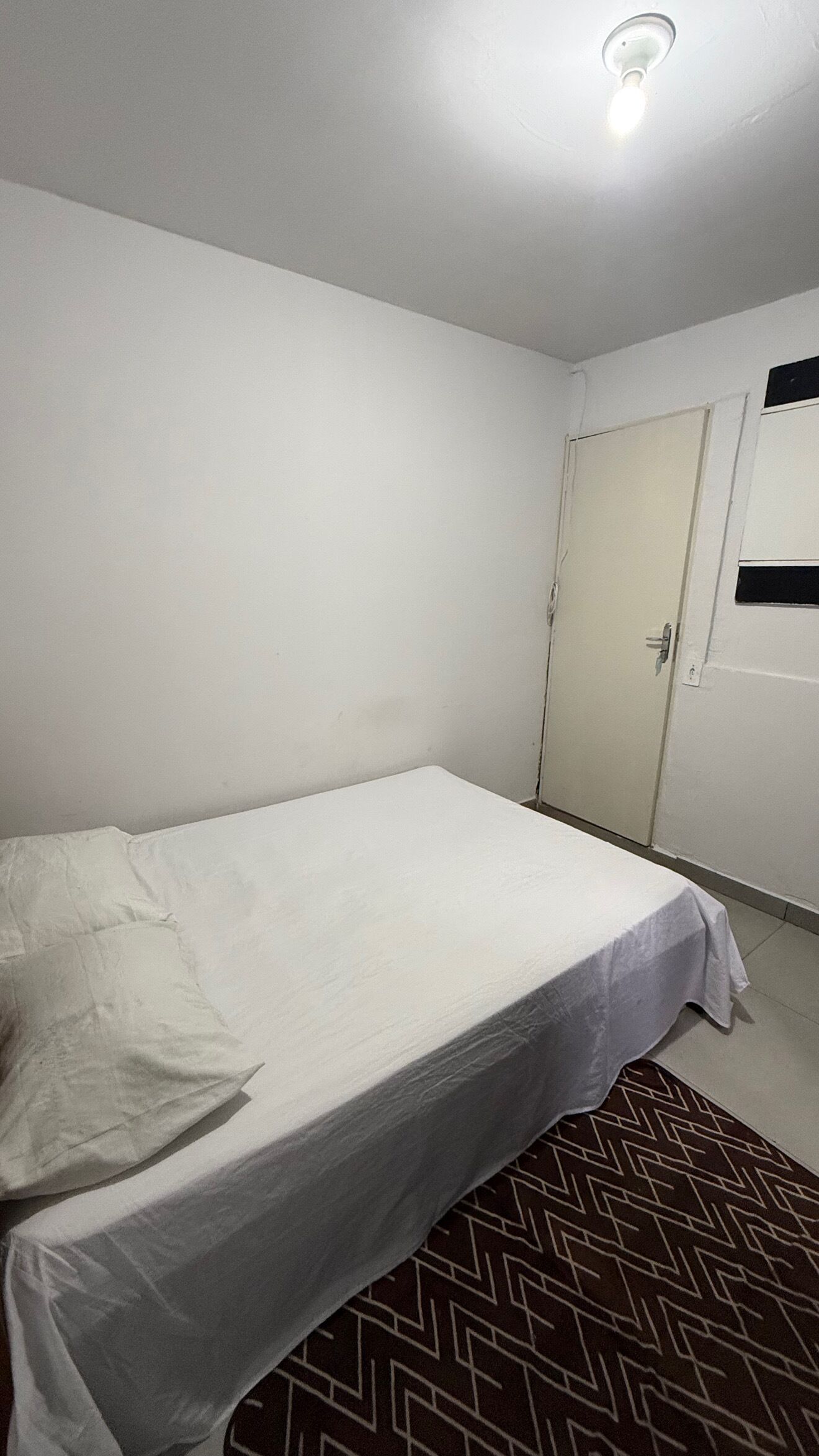 Een eenpersoonskamer met een simpel wit bed, een deur met een raam en een kamerbreed tapijt met dessin.
