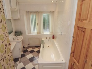 A bathroom with a checkered floor, a bathtub, a sink, a window with curtains, and a wooden door. - The Haven (Morecambe)