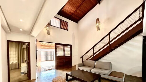 A spacious living area with a sofa, coffee table, and wicker ottomans. A staircase with a wooden railing leads to another level.