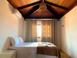 A bedroom with a wooden ceiling, a bed with white bedding, a nightstand, a window with curtains, and a hanging light fixture.
